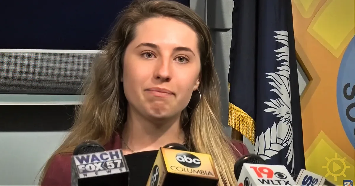 A woman with long hair stands behind microphones with "WACH FOX 57" and "ABC Columbia" logos, in front of a flag.