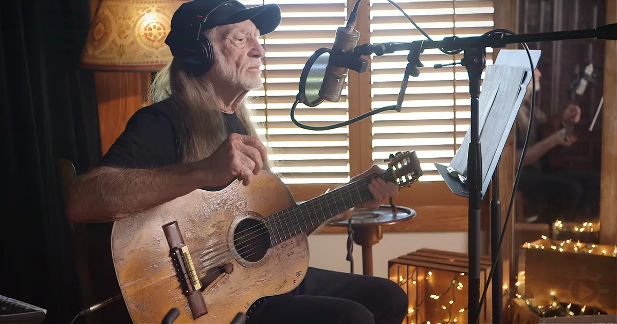 An older man with long hair and a beard plays an acoustic guitar in a warmly lit room.
