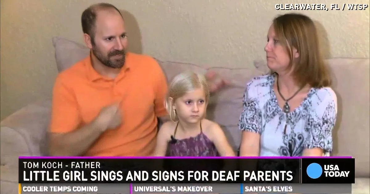 A family of three sits on a couch, with a man in an orange shirt gesturing, while a woman and a young girl listen.