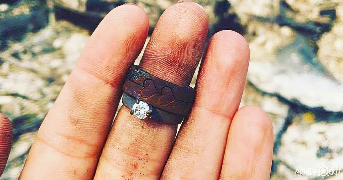 A hand holds a ring with a heart design and a central diamond, set against a blurred natural background.