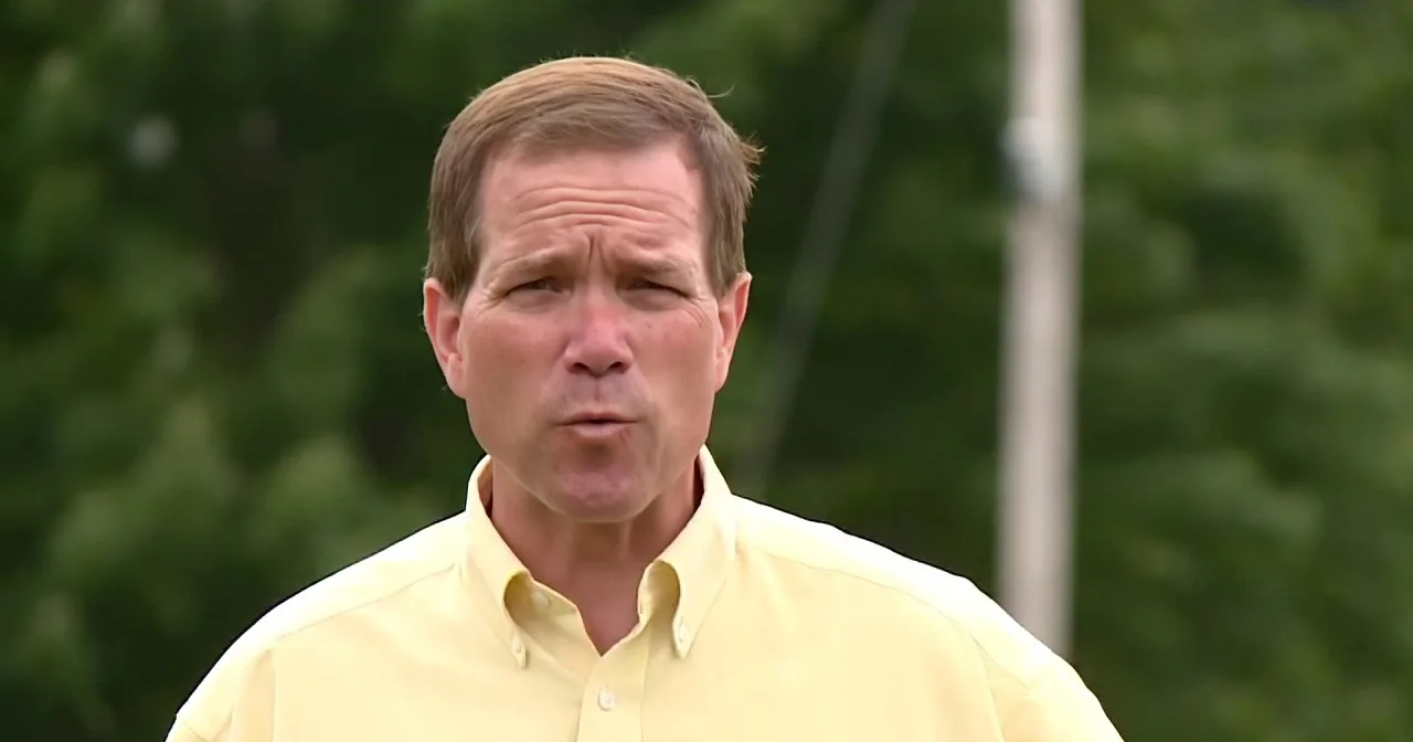A man in a yellow shirt stands outdoors, looking slightly to the side with a serious expression.