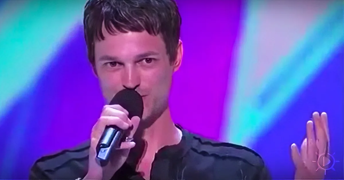 A person holding a microphone, wearing a black shirt, with colorful stage lights in the background.