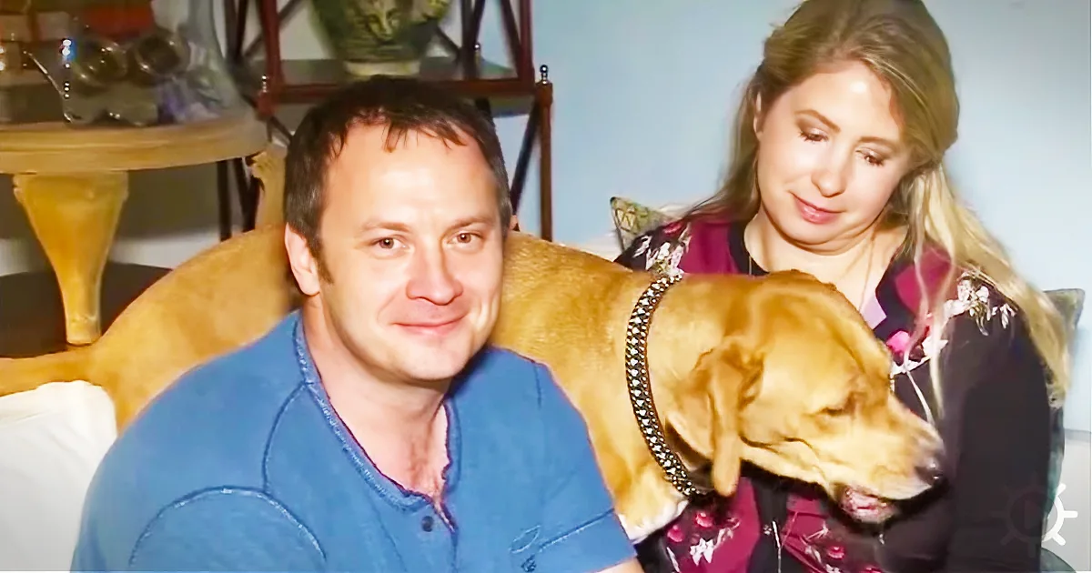 A man in a blue shirt smiles as a dog rests on his shoulder, with a woman in the background.