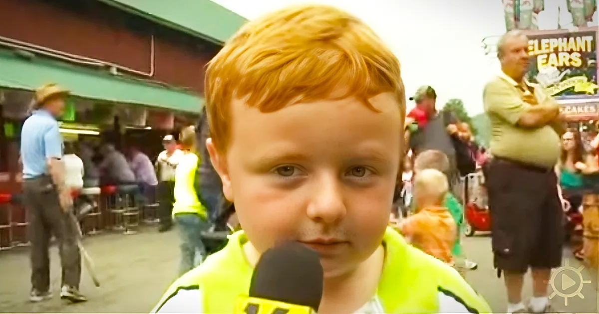 A young boy with red hair, wearing a yellow shirt, speaks into a microphone in a crowded outdoor setting.