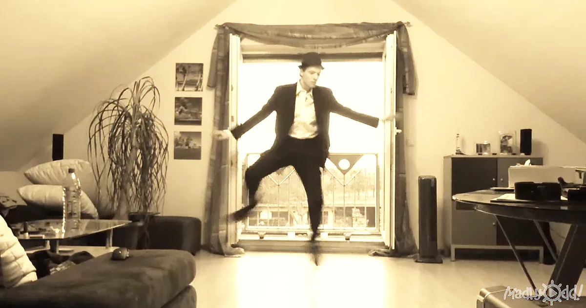 A man in a suit and hat dances in a room with a slanted ceiling and a large window.