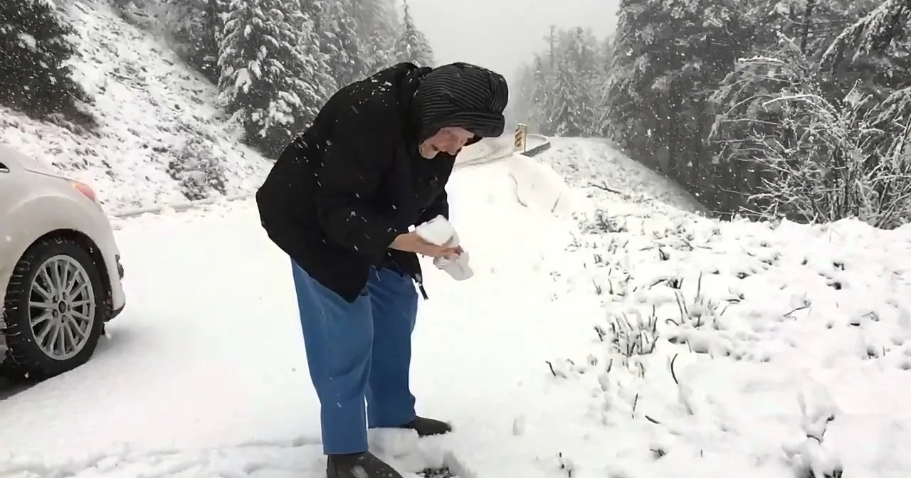 A person in winter clothing bends over in the snow, holding a snowball near a car on a snowy road.