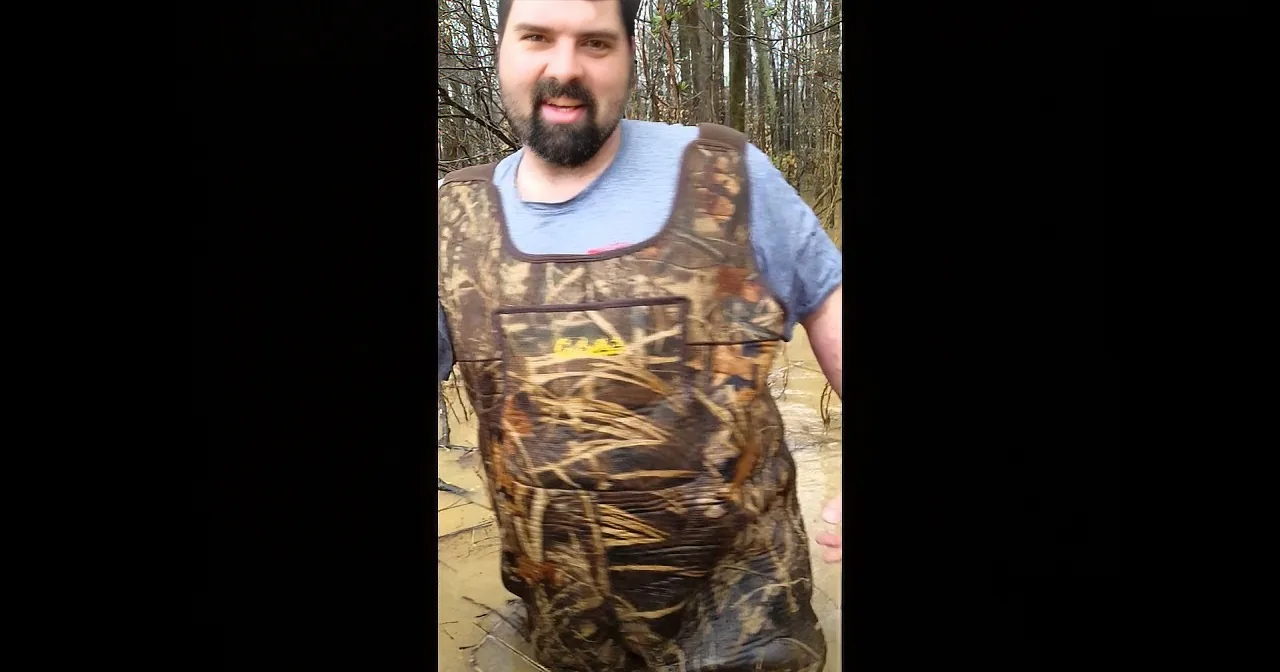 A man in camouflage hunting gear stands in a wooded area, smiling.