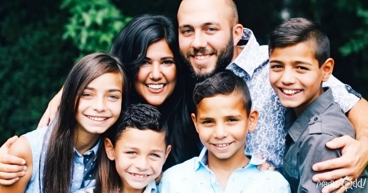A family of five smiles together outdoors, with a lush green background.