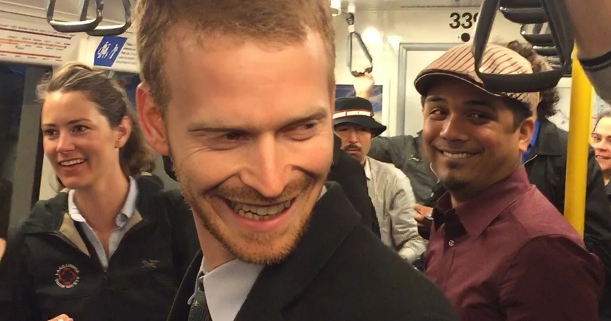 A man with a beard and short hair smiles widely on a crowded subway, surrounded by other passengers.