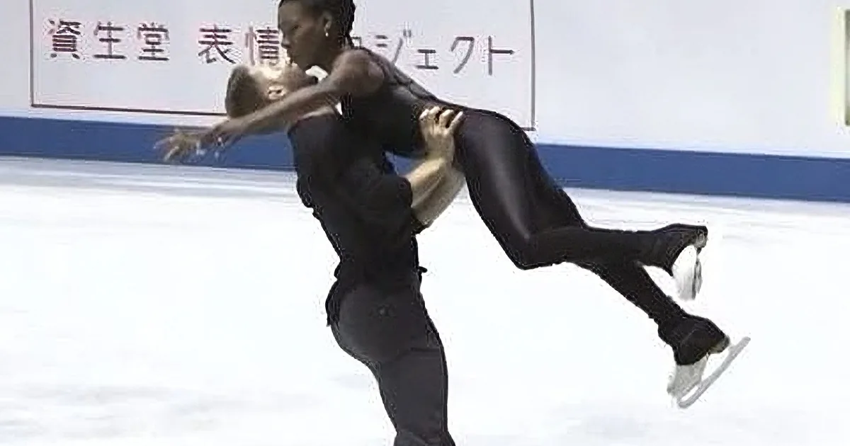 A figure skater in a black outfit lifts another skater in a dramatic pose on the ice.