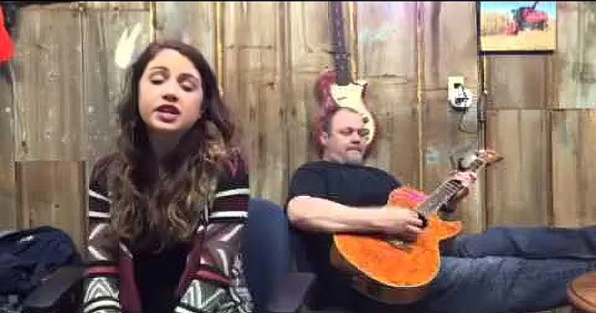 A woman sings while a man plays guitar in a rustic setting with wooden walls.
