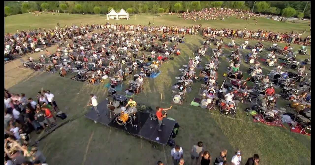 A large crowd gathers around a massive drum circle in a grassy field.