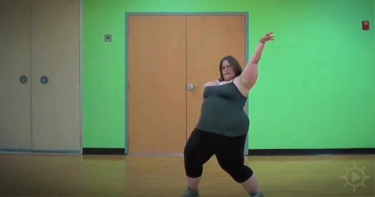 A person in a green tank top and black leggings is dancing in a room with green walls and wooden floors.