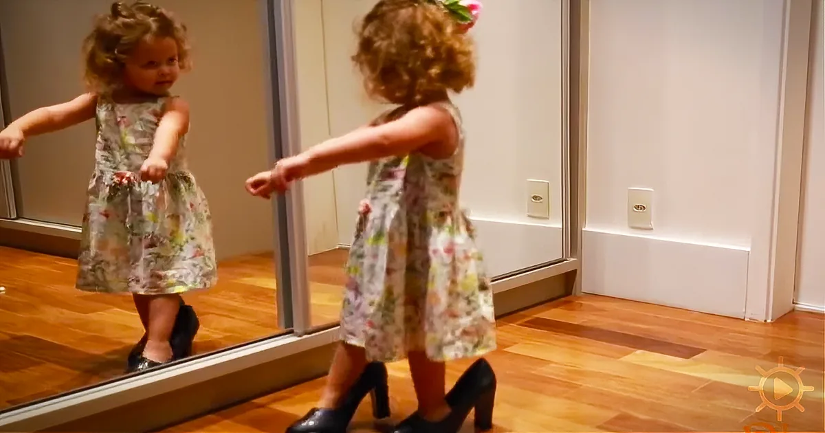 A young girl in a floral dress and high heels stands in front of a mirror, arms outstretched.