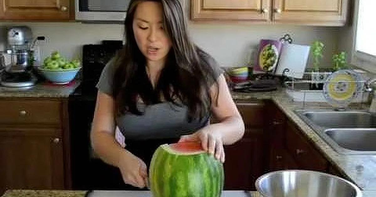 A woman in a kitchen slices a watermelon on a cutting board.