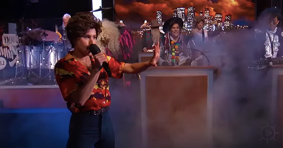 A woman in a colorful shirt sings into a microphone on stage, with a cityscape backdrop and musicians behind her.