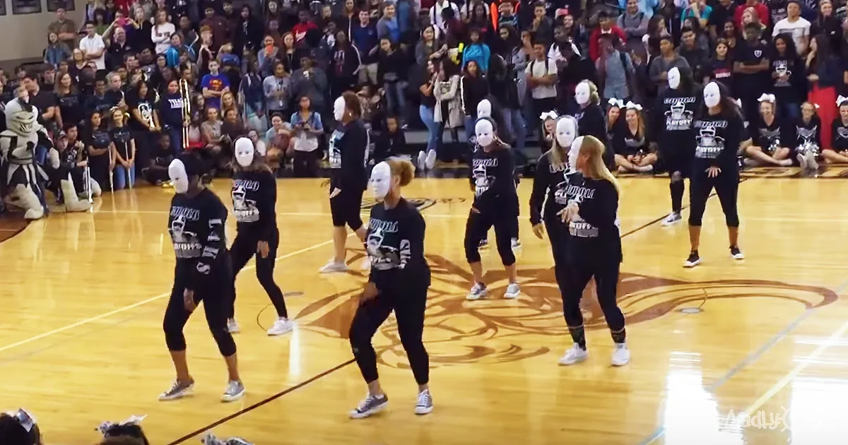 A group of dancers in black outfits and white masks perform on a gymnasium floor, watched by a cheering crowd.