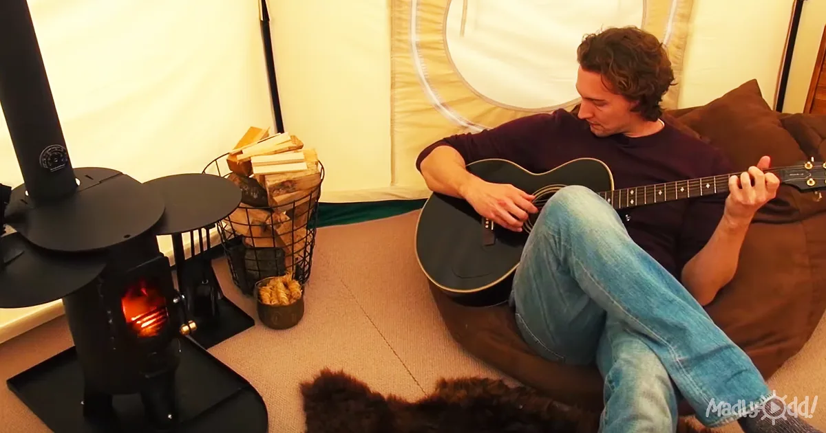 A man sits on a couch playing guitar by a lit wood stove in a cozy tent setting.