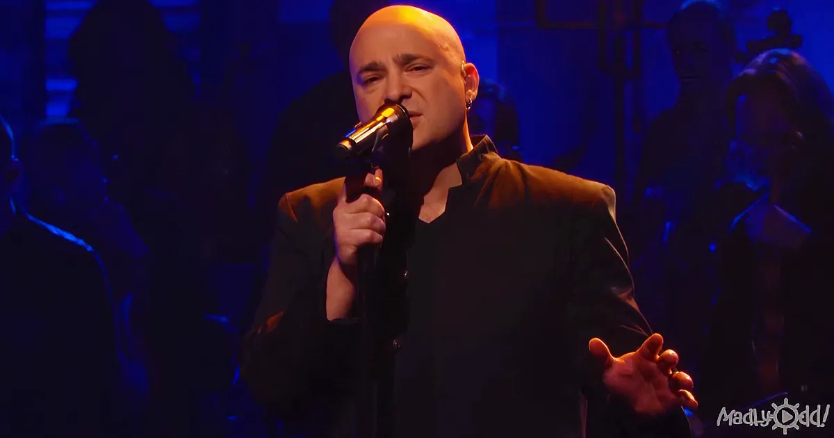 A man in a black suit sings into a microphone on stage with a blue-lit background.