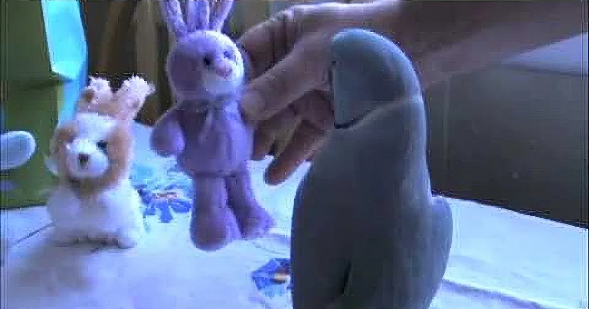 A hand holds a purple stuffed rabbit, surrounded by other stuffed animals on a table.
