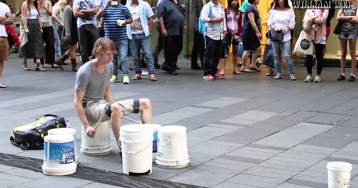 A street performer plays drums with buckets on a busy city sidewalk, surrounded by onlookers.