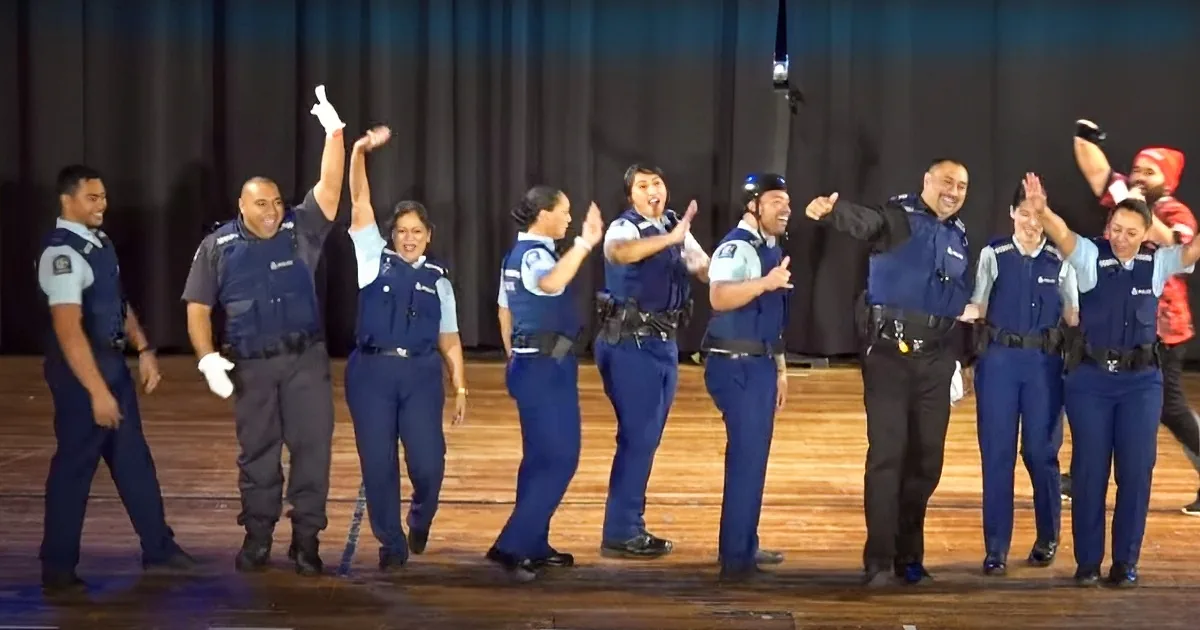 Police officers in blue uniforms are dancing and smiling on a stage.