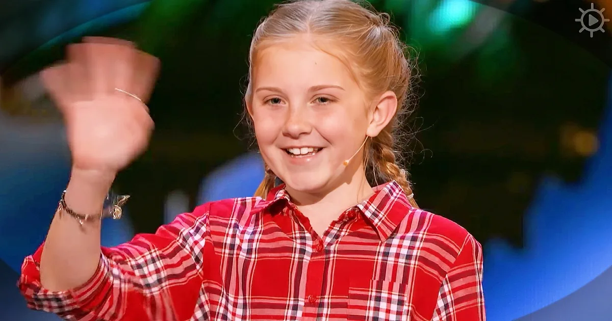 A girl in a red plaid shirt smiles while waving, wearing a microphone and a bracelet.