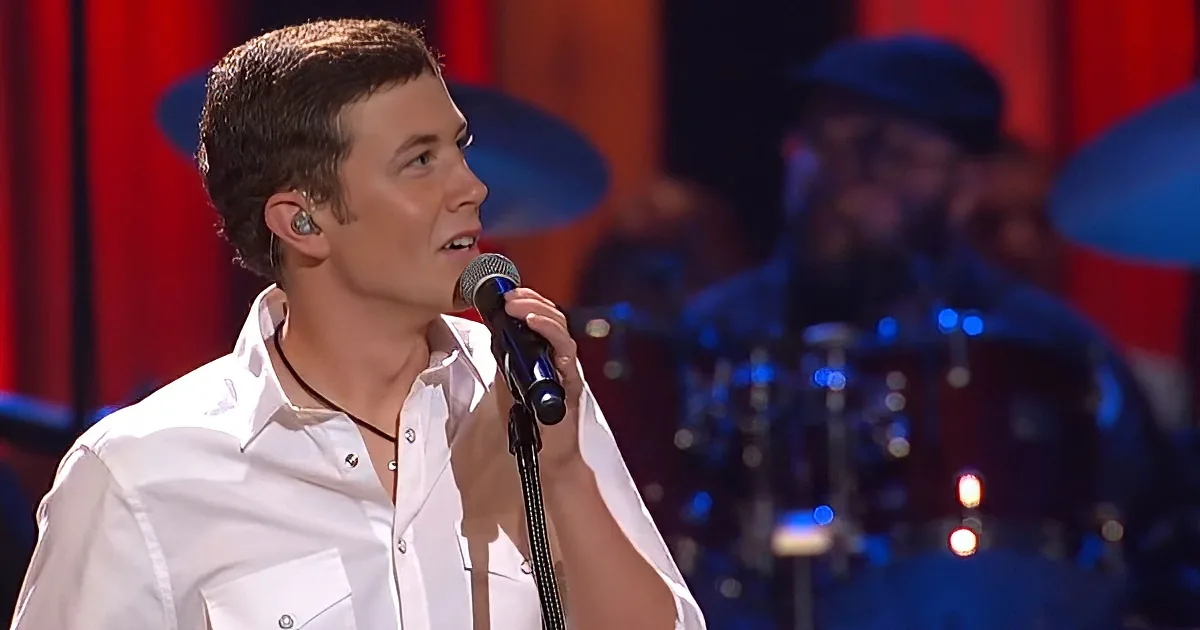 A man in a white shirt sings into a microphone, wearing an earpiece, with a drummer in the background.