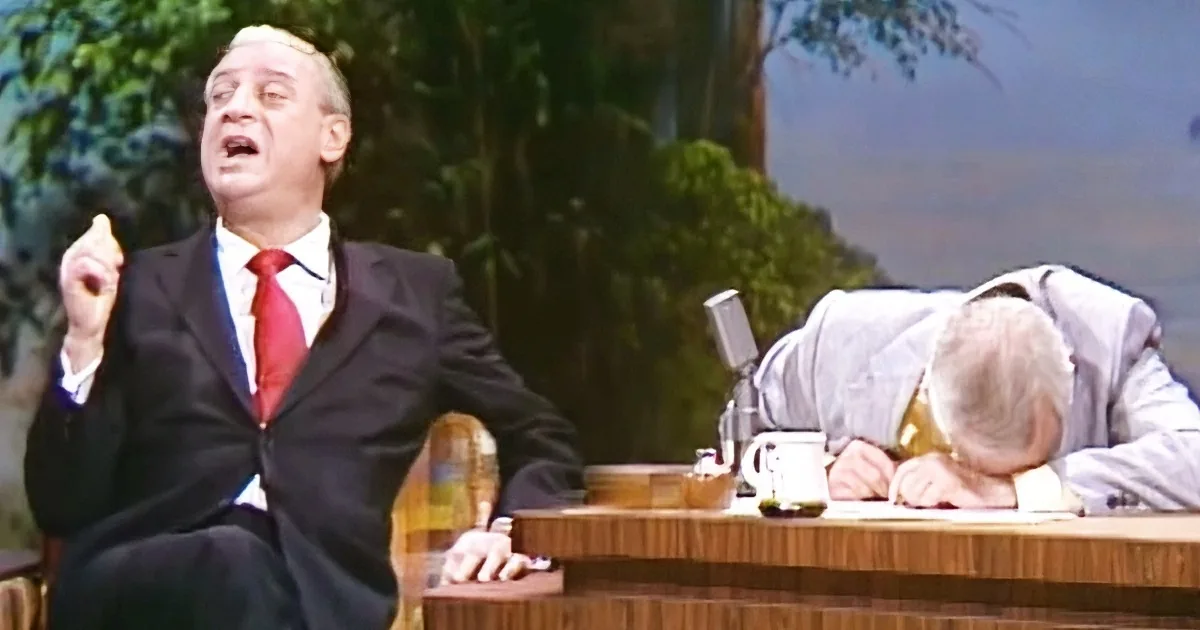 A man in a suit and tie is gesturing passionately while another man sleeps on a desk.