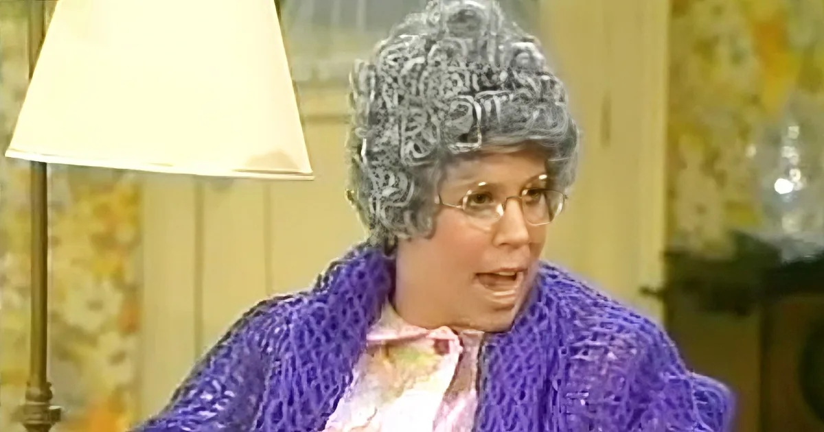 A person with curly gray hair and glasses, wearing a purple sweater, is speaking in a room with a lamp and floral wallpaper.