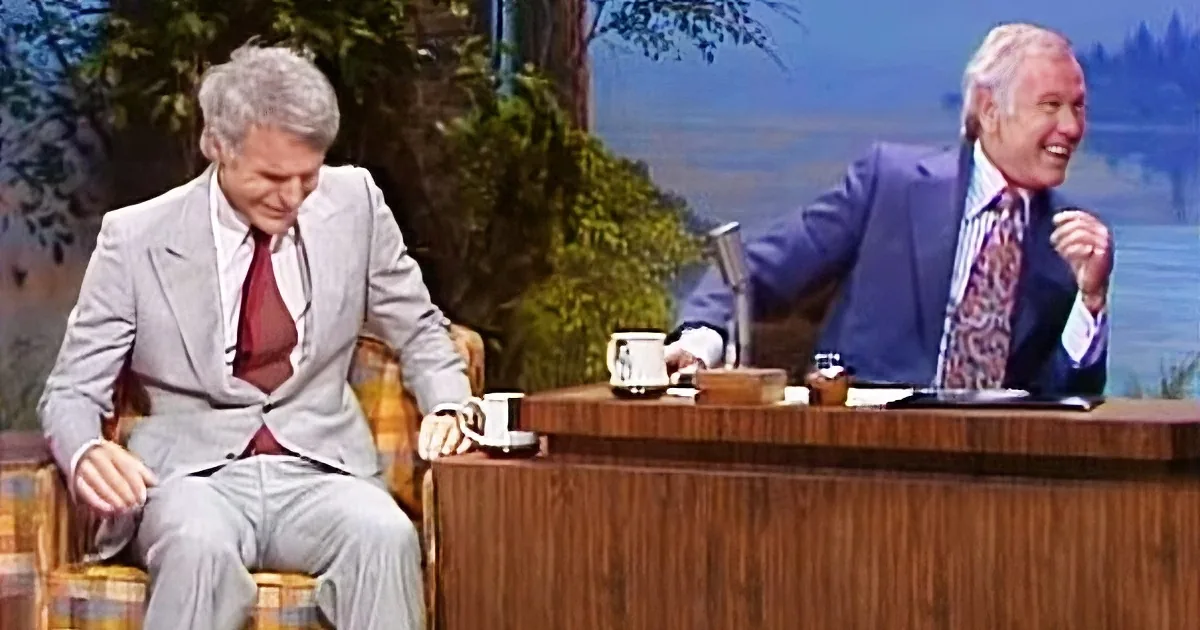 A man in a suit sits on a chair, laughing, with another man behind a desk also laughing.