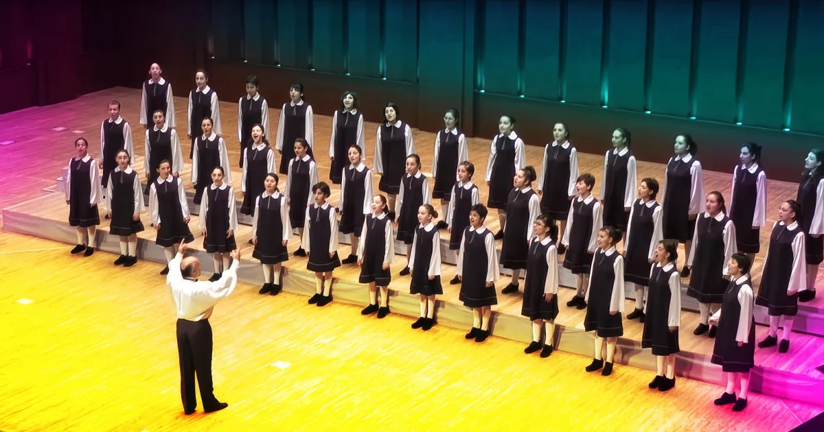 A choir of young girls in black and white uniforms stands in formation on a stage.