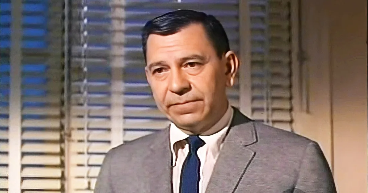A man in a gray suit and blue tie stands in front of a window with blinds.