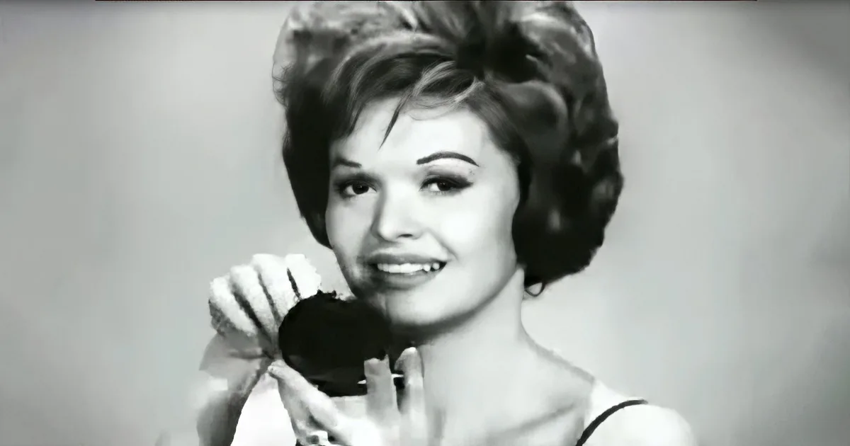 A woman with short, styled hair smiles while holding a black object near her face.