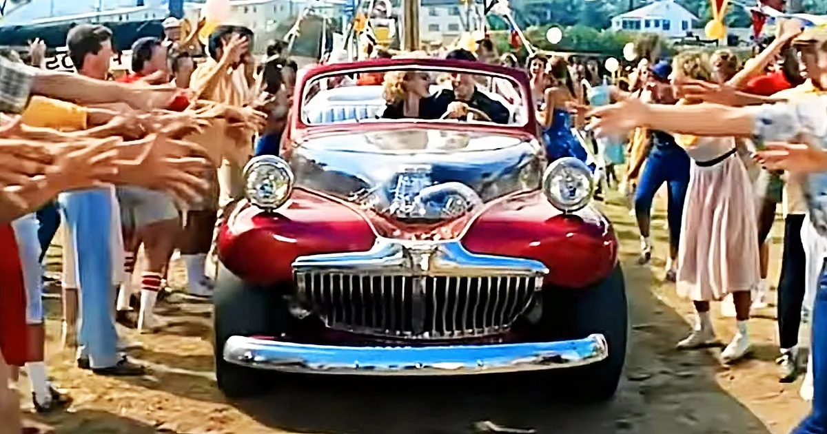 A red vintage car drives through a crowd of people waving and cheering.