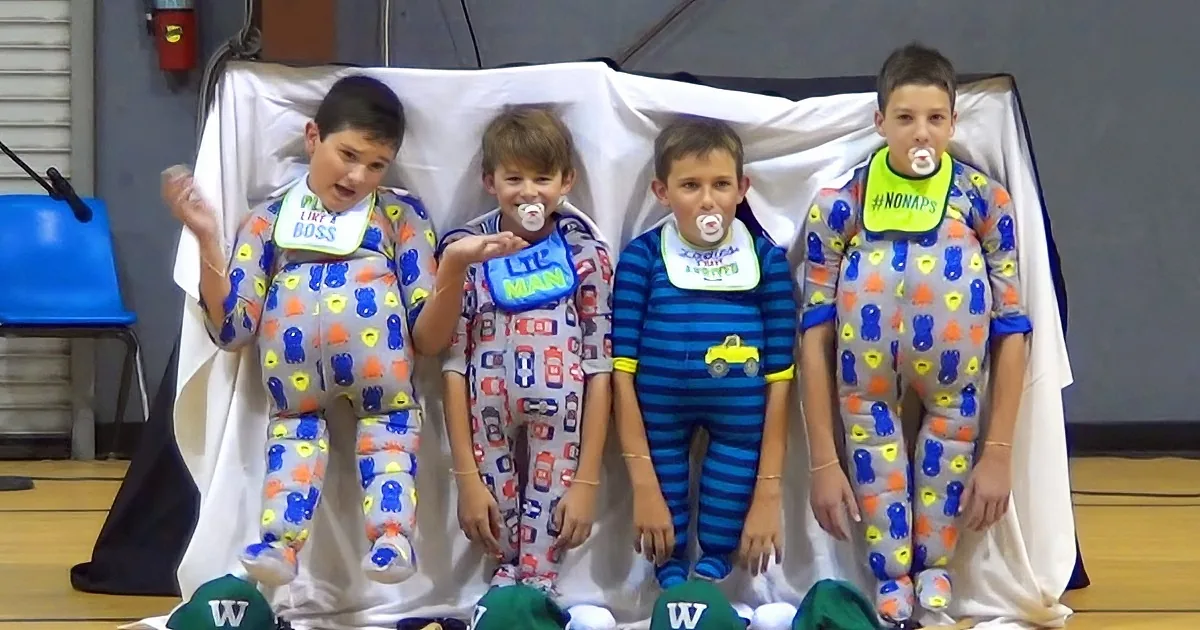 Four boys in pajamas with pacifiers and bibs stand on a stage, smiling.