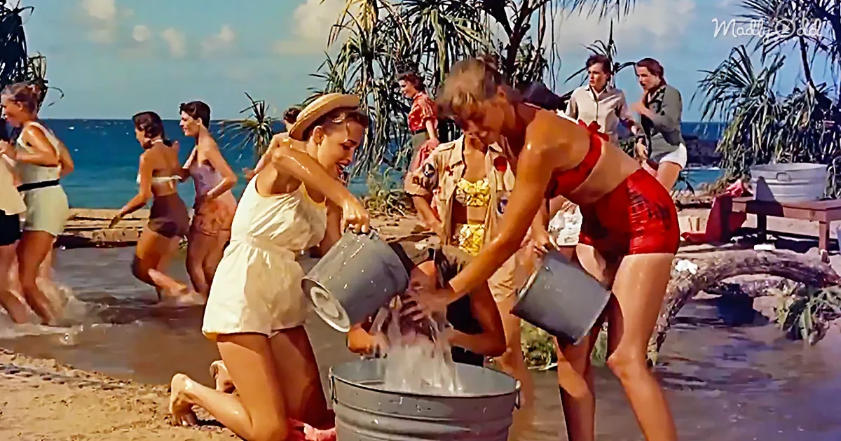 Two women pour water from buckets into a metal bucket on a beach.