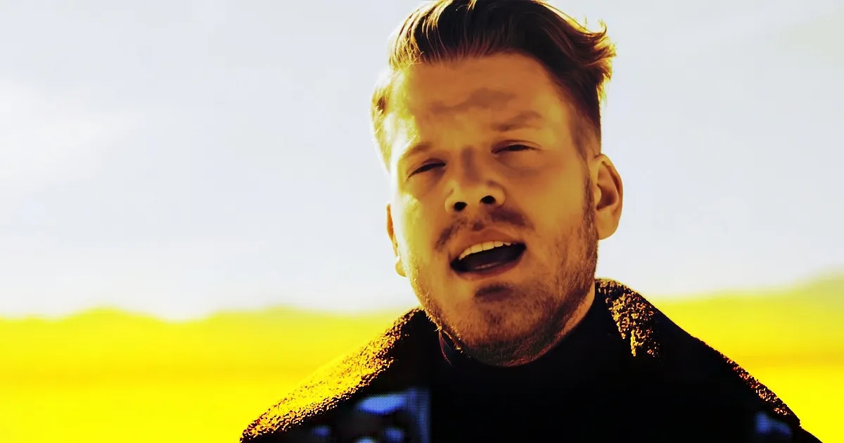 A man with short, styled hair and a beard, wearing a dark jacket, stands against a bright, yellow-hued background.