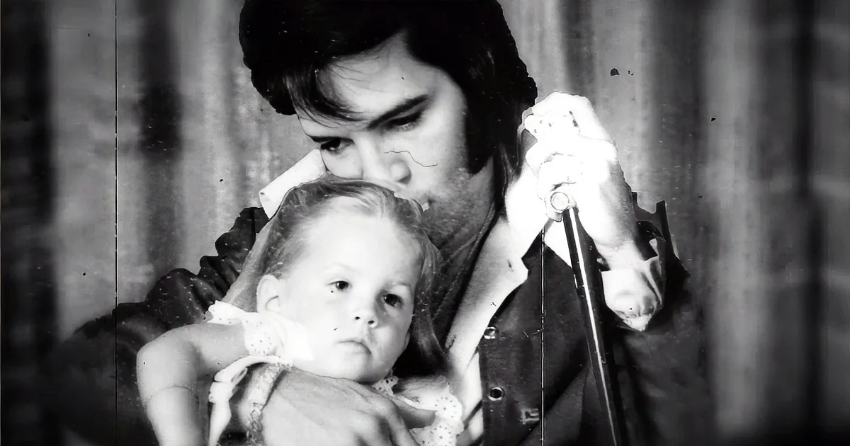 A man holds a baby, both looking forward, with a curtain in the background.