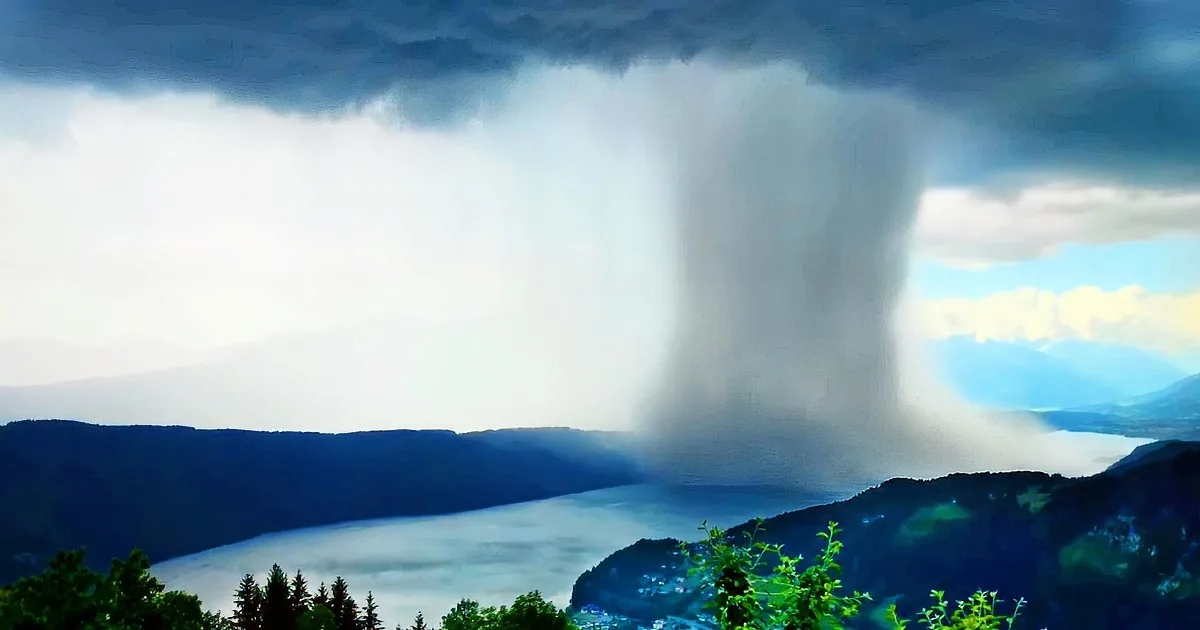 A massive, dark cloud pours rain over a serene landscape with mountains and a lake.