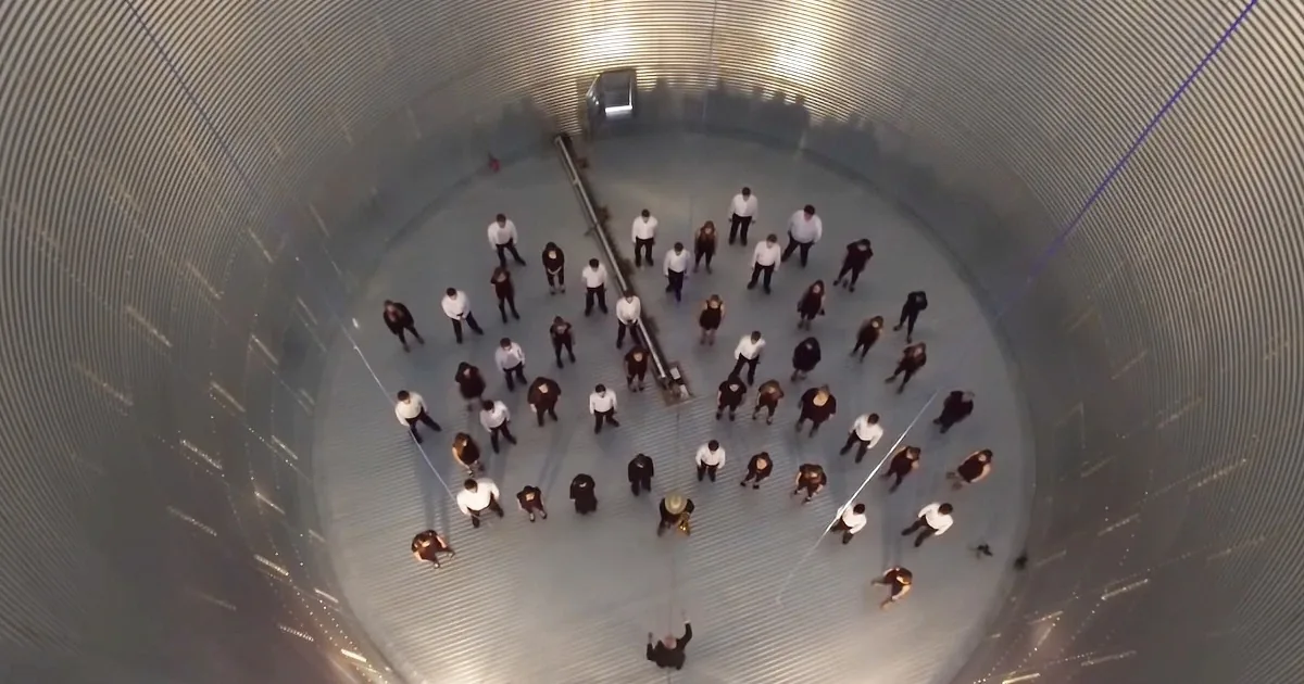 A group of people in white shirts and black pants are arranged in a circle inside a large, cylindrical structure with...