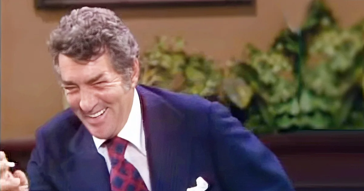A man in a blue suit and red tie is smiling and leaning forward, with green plants in the background.