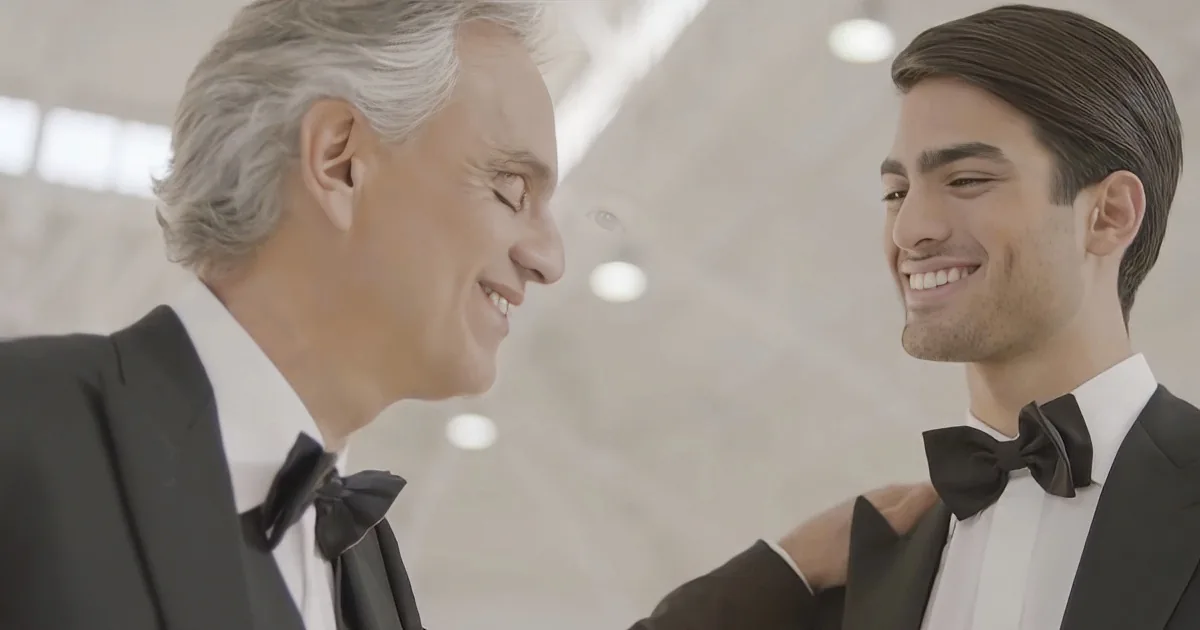 Two men in tuxedos smile at each other, one older with gray hair, the other younger with dark hair.
