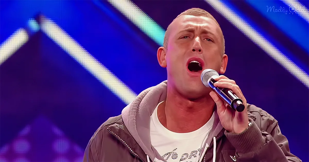 A man sings passionately into a microphone, wearing a hoodie and white shirt, against a blue stage backdrop.