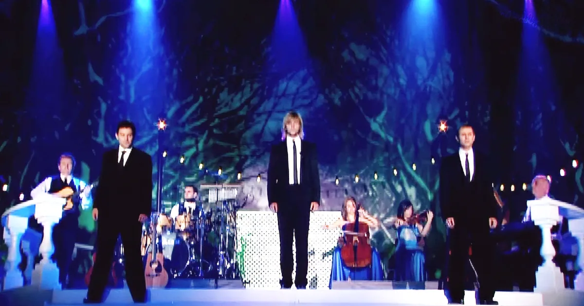 Three men in suits stand on stage with musicians and a forest backdrop.