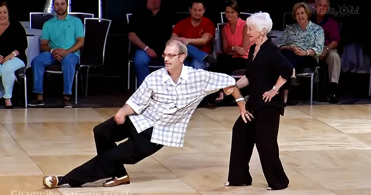An older man in a checkered shirt and black pants is dancing with a woman in black on a wooden floor, with spectators in...
