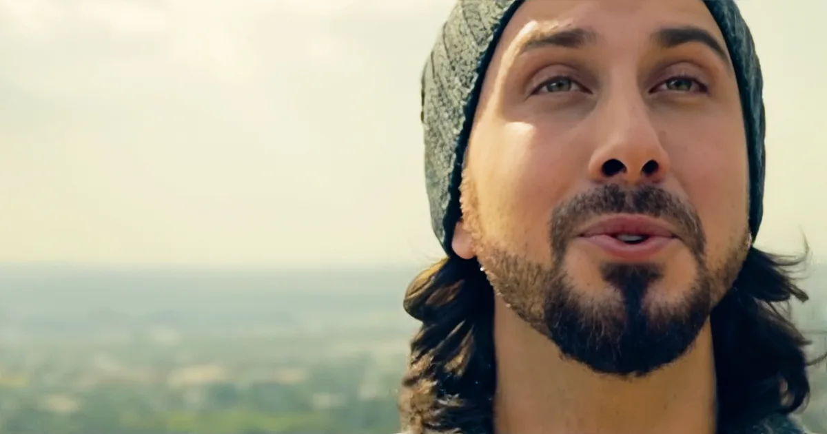 A man with long hair and a beard wearing a beanie, looking upward with a blurred landscape in the background.