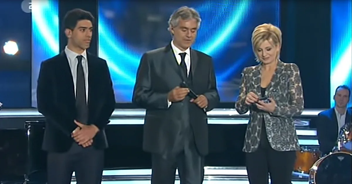 A man in a suit holds a microphone on stage with two others, one in a patterned blazer.