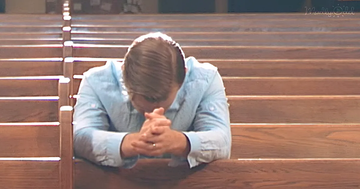 A man in a light blue shirt is sitting on a wooden pew, hands clasped, head bowed in prayer.