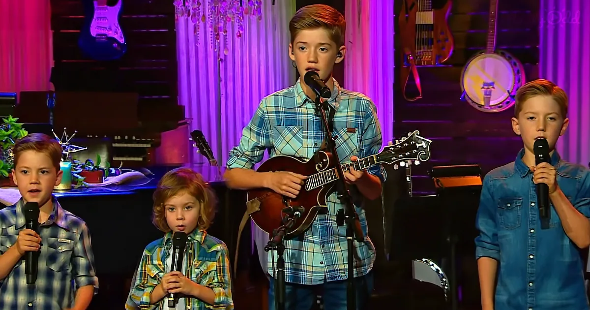A young boy in a plaid shirt plays a mandolin while singing, accompanied by three other children holding microphones.